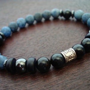 May include: A bracelet with a mix of blue, black, and silver beads. It features matte blue-grey beads, glossy black beads, and a decorative silver-toned cylindrical bead. The bracelet is strung on an elastic cord. The beads are approximately 0.8 cm in diameter.
