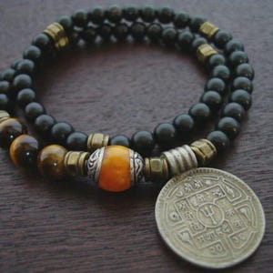 May include: A layered bracelet featuring black beads, tiger eye beads, and gold-toned metal accents. An orange and silver bead is centered on the bracelet. A silver coin with engraved text rests beside the bracelet.