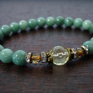 May include: A beaded bracelet featuring round, light green jade beads. The bracelet has a central decorative element with a faceted yellow crystal, accented with small clear crystals and gold-toned metal.