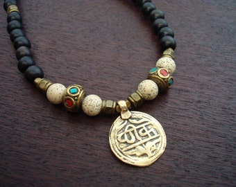 May include: A beaded necklace featuring black wooden beads, speckled white beads, and brass accents. The necklace includes a gold-colored pendant with engraved text and colorful stone embellishments.