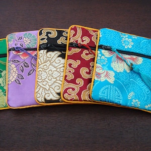 May include: Five decorative fabric pouches in various colours, each with a floral design. The pouches are green, purple, black, red, and turquoise, with gold trim and a zip. The turquoise pouch has a tassel.
