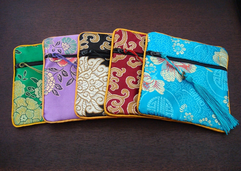 May include: Five decorative fabric pouches in various colours, each with a floral design. The pouches are green, purple, black, red, and turquoise, with gold trim and a zip. The turquoise pouch has a tassel.