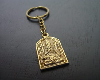 May include: A gold-colored metal keychain featuring a detailed relief of a seated deity within an arched frame. The keychain includes a ring and chain, and the deity is adorned with intricate details. The background is a dark surface.
