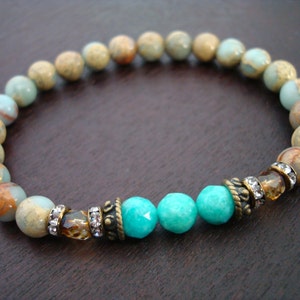 Könnte beinhalten: Ein Armband mit einer Mischung aus erd- und türkisfarbenen Perlen. Das Armband enthält dekorative Metallakzente mit klaren Kristallverzierungen, ein stilvolles Accessoire.