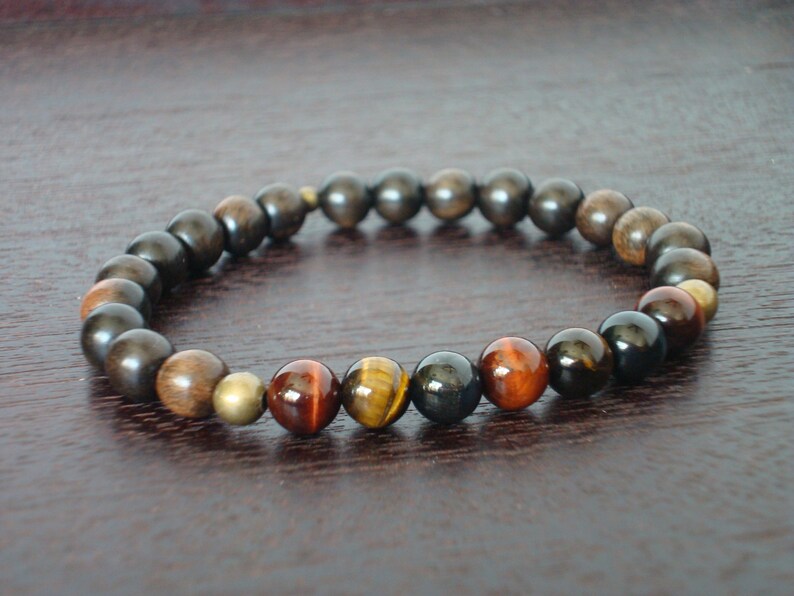 May include: A bracelet made of wooden and gemstone beads. The bracelet features dark brown, light brown, and golden beads, with tiger's eye and brass beads. The bracelet is strung on an elastic band. The beads are approximately 0.8 cm in diameter.