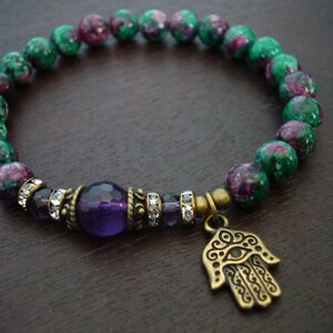 May include: A green and purple gemstone bracelet with a purple amethyst bead and a hamsa hand charm. The bracelet is made with a stretchy cord and is approximately 7 inches in length.
