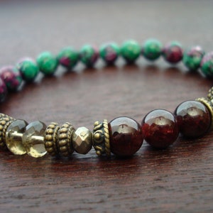 May include: A bracelet made with green and pink gemstone beads, brown and gold metal beads, and dark red gemstone beads.