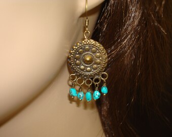 May include: A brass-colored earring with a circular design featuring a central stud and surrounding dots. Small turquoise beads dangle from the bottom. The earring is suspended from a gold-colored hook.