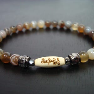 Könnte beinhalten: Armband mit Perlen und einer zentralen cremefarbenen Perle mit Gravur. Das Armband hat eine Mischung aus braunen, beigen und weißen Perlen, getrennt durch silberfarbene Metallakzente. Die Perlen sind auf einer flexiblen Schnur aufgefädelt.
