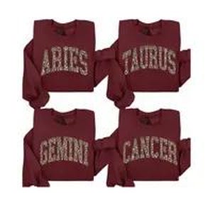 May include: Four maroon sweatshirts, each displaying a different zodiac sign in a gold, leopard-print style font: Aries, Taurus, Gemini, and Cancer. The sweatshirts have long sleeves and a crew neck.