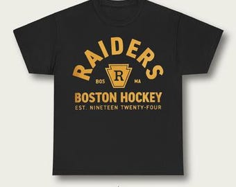 May include: Black t-shirt with a gold arched "RAIDERS" design above a shield logo and "BOSTON HOCKEY" text. The text "EST. NINETEEN TWENTY-FOUR" is below. The shirt is a classic crew neck style.
