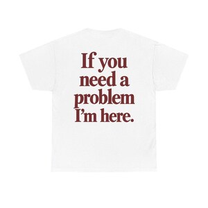 May include: A white short-sleeved t-shirt featuring dark red text printed on the back. The text reads, "If you need a problem I'm here." The shirt has a classic crew neck design and is made of a plain, solid-colored fabric with no other visible patterns.