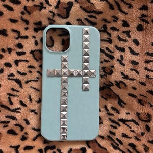 Puede incluir: Funda de teléfono azul claro con un diseño de cruz tachonada plateada. La funda tiene forma rectangular con esquinas redondeadas y un recorte para la cámara. La cruz tachonada está centrada.