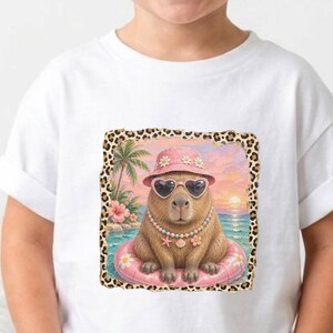 May include: White t-shirt featuring a cartoon capybara wearing sunglasses, a pink hat, and a necklace, floating on a pink inner tube. The capybara is set against a tropical beach scene with palm trees and a sunset.