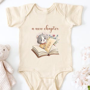 May include: A cream-colored baby onesie with the text "a new chapter" above a watercolor illustration of a gray and white animal reading a book. A teddy bear and flowers are in the background.