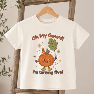 May include: A natural-colored short-sleeve t-shirt featuring a graphic of a smiling pumpkin wearing a party hat and holding a green leaf balloon. The text above reads Oh My Gourd! and the text below reads I'm turning five! in brown lettering.