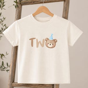May include: A natural-colored short-sleeve t-shirt featuring a graphic print of a brown bear wearing a blue polka-dot party hat. The text "TWO" is printed in large, light brown letters next to the bear illustration on the front of the shirt.