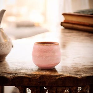 Könnte beinhalten: Eine kleine, runde, rosa Keramiktasse mit strukturierter Oberfläche und braunem Rand. Die Tasse steht auf einem dunklen Holztisch. Die Tasse ist wahrscheinlich für Tee oder Kaffee.
