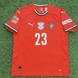 Peut inclure: Un maillot de football rouge avec le numéro 23 en blanc. Le maillot présente des bordures blanches et noires sur le col et les manches. Des logos et des écussons sont visibles sur la poitrine et les manches. Le tissu est respirant.