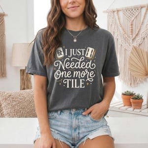 May include: Dark gray t-shirt with the text "I Just Needed One More Tile" in a whimsical font. The design includes illustrations of Mahjong tiles. The shirt is a casual, comfortable style.
