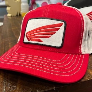 May include: A red trucker hat with a white mesh back and a red and white embroidered patch. The patch features a stylized wing design. The hat has a curved brim with white stitching.