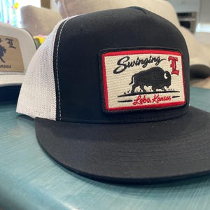 May include: Black and white trucker hat with a flat brim. The hat features a rectangular patch with the text "Swinging I" above a bison graphic and "Lebo, Kansas" below. A second hat is visible in the background.