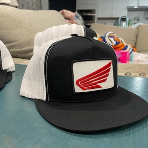 May include: Black and white trucker hat with a black brim and a white mesh back. The hat features a rectangular patch with a red wing design. The hat is sitting on a blue surface.