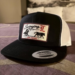 Op de afbeelding: Zwarte en witte truckerpet met een platte klep. Het voorpaneel is zwart met een rechthoekige patch met een cowboy die een stier vastbindt en de tekst "Swinging L Lebo, Kansas". De achterkant is wit mesh.
