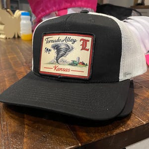 以下が含まれることがあります： 「Tornado Alley Kansas」と書かれたパッチが付いた、黒と白のトラッカーハット。 パッチには竜巻のグラフィック、農場のシーン、文字「L」が描かれています。 帽子は黒いつばと白いメッシュの背面を備えています。