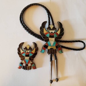 May include: A Native American bolo tie featuring a multi-colored geometric design with a black leather cord. The bolo tie is accompanied by a matching pin.