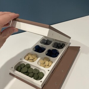 May include: A brown, book-shaped storage container with a white interior featuring eight separate compartments. The compartments are filled with small, smooth, pebble-like pieces in shades of dark gray, navy blue, cream, and olive green.