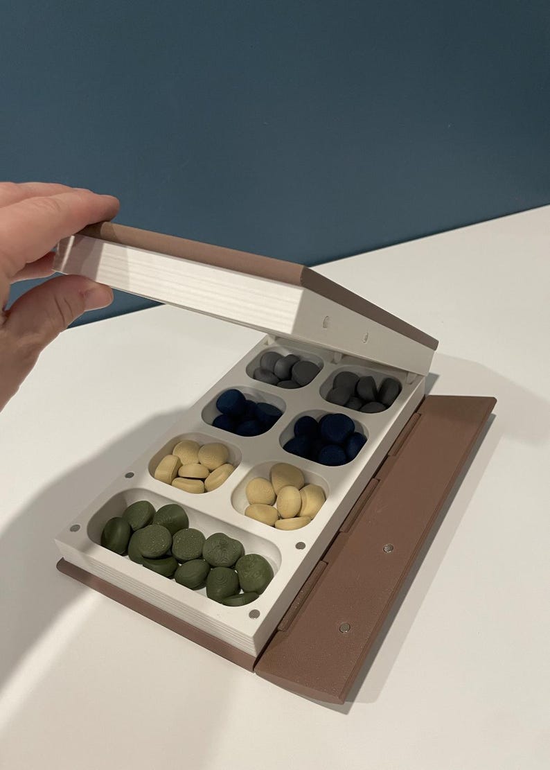 May include: A brown, book-shaped storage container with a white interior featuring eight separate compartments. The compartments are filled with small, smooth, pebble-like pieces in shades of dark gray, navy blue, cream, and olive green.