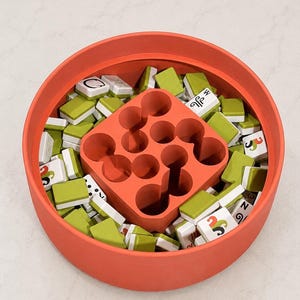 Puede incluir: Un recipiente redondo de color coral lleno de numerosas fichas de juego rectangulares peque&ntilde;as con reverso verde y caras blancas con varios s&iacute;mbolos negros y de colores. Un inserto cuadrado de color coral con nueve huecos circulares en el centro.