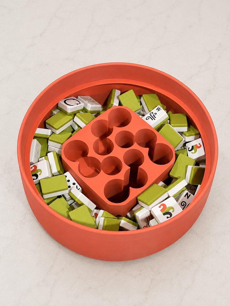 Puede incluir: Un recipiente redondo de color coral lleno de numerosas fichas de juego rectangulares peque&ntilde;as con reverso verde y caras blancas con varios s&iacute;mbolos negros y de colores. Un inserto cuadrado de color coral con nueve huecos circulares en el centro.