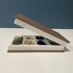 May include: A rectangular storage container shaped like a closed book with a brown cover and white pages. The top lid is propped open to reveal a white interior tray divided into six compartments, each filled with small, smooth, round tokens in green, tan, dark blue, and gray.