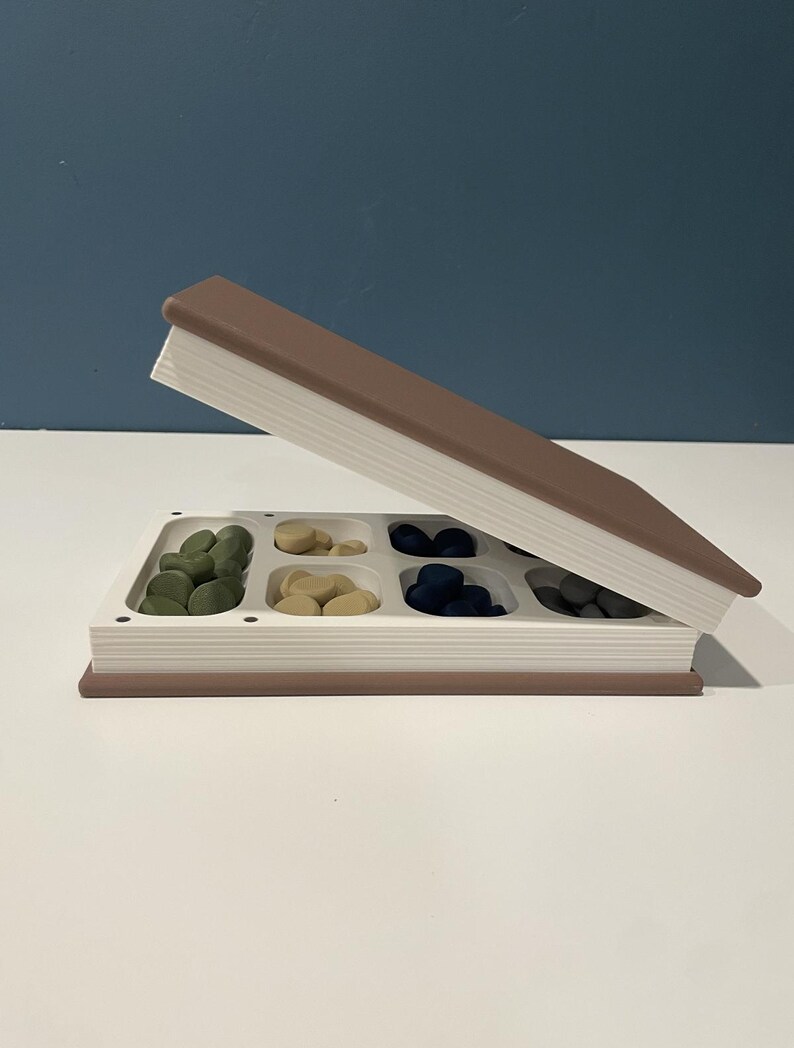 May include: A rectangular storage container shaped like a closed book with a brown cover and white pages. The top lid is propped open to reveal a white interior tray divided into six compartments, each filled with small, smooth, round tokens in green, tan, dark blue, and gray.