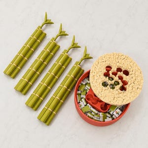 Puede incluir: Un juego de mesa que incluye cuatro soportes de pl&aacute;stico verde con forma de bamb&uacute; y un recipiente redondo rojo con una tapa beige texturizada. El recipiente contiene fichas rectangulares peque&ntilde;as con marcas verdes y blancas y una bandeja.