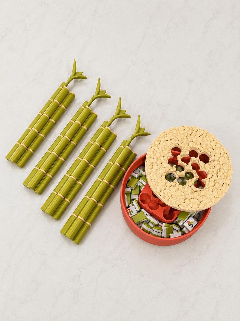 Puede incluir: Un juego de mesa que incluye cuatro soportes de pl&aacute;stico verde con forma de bamb&uacute; y un recipiente redondo rojo con una tapa beige texturizada. El recipiente contiene fichas rectangulares peque&ntilde;as con marcas verdes y blancas y una bandeja.