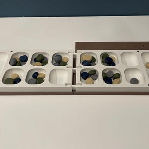 May include: A foldable board game with a white plastic frame featuring twelve square recessed pits and two elongated end storage areas. The board contains small, smooth, rounded game pieces in shades of blue, green, gray, and tan.