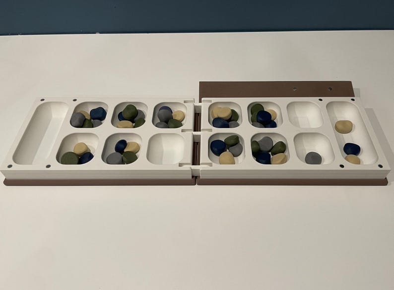 May include: A foldable board game with a white plastic frame featuring twelve square recessed pits and two elongated end storage areas. The board contains small, smooth, rounded game pieces in shades of blue, green, gray, and tan.