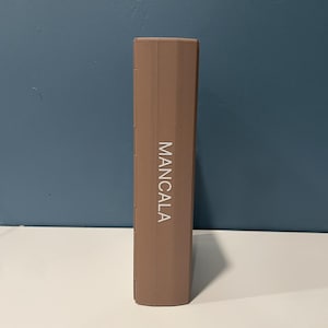 May include: A rectangular, light brown, closed Mancala game board standing vertically. The word MANCALA is printed in white, capital letters vertically along the center of the front panel. The object has a smooth, solid-colored exterior surface.