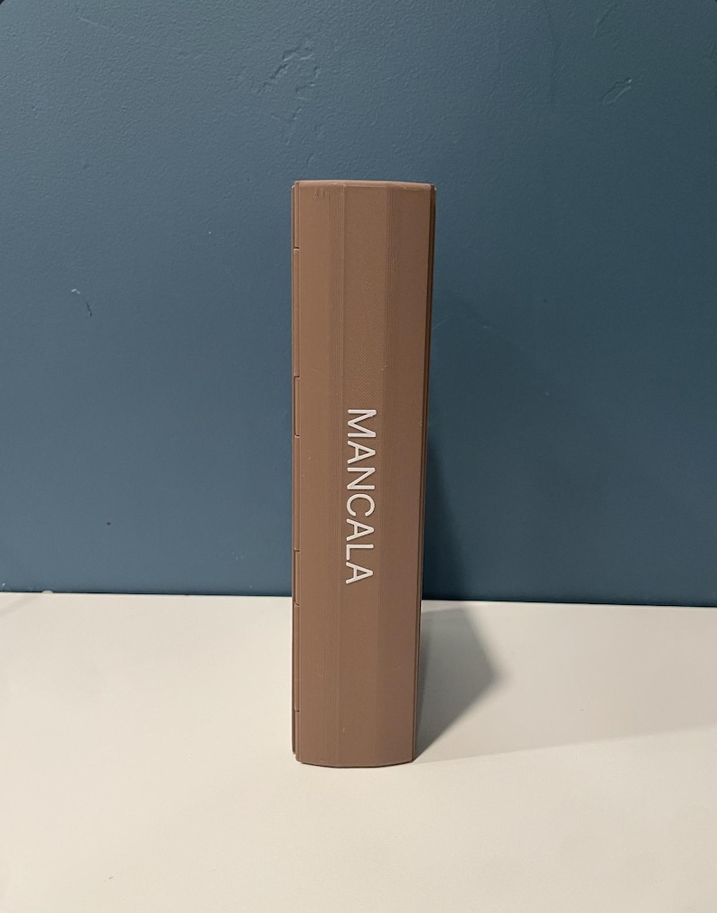 May include: A rectangular, light brown, closed Mancala game board standing vertically. The word MANCALA is printed in white, capital letters vertically along the center of the front panel. The object has a smooth, solid-colored exterior surface.