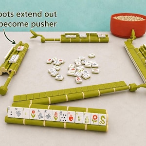 Puede incluir: Un juego de Mahjong con cuatro soportes verdes estilo bamb&uacute; con brazos empujadores extensibles. Los soportes sostienen fichas blancas con varios s&iacute;mbolos, n&uacute;meros y dise&ntilde;os florales. Un mont&oacute;n de fichas sueltas se encuentra en el centro.