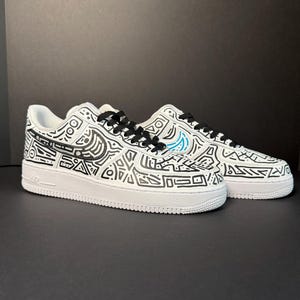 May include: White trainers with black abstract patterns and black laces. The shoes feature a blue swoosh logo and are set against a dark background. The trainers are a popular style.