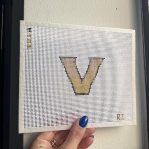 May include: A white canvas with a hand-stitched letter "V" in beige and yellow thread. The canvas has a grid-like texture and the initials "RI" are stitched in the bottom right corner. The canvas is held by a hand with blue nail polish.