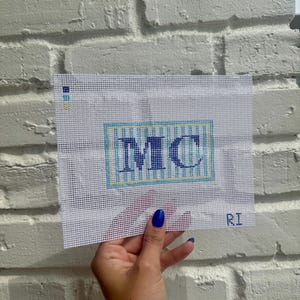 May include: A needlepoint canvas featuring the letters "MC" in a blue and white striped design, framed by a light blue border. The canvas is held against a white brick wall. The initials "RI" are visible in the bottom right corner.