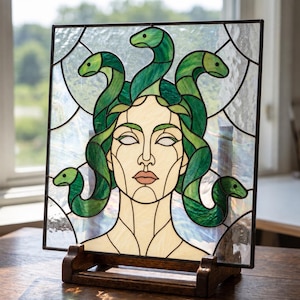 May include: A square stained glass panel featuring a central figure with a pale face and dark outlines, surrounded by multiple green snakes. The background consists of clear, textured glass pieces held together by dark metal lead lines.
