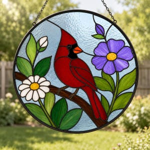 May include: A circular stained glass suncatcher featuring a red cardinal perched on a brown branch. The design includes a white daisy, a purple flower, green leaves, and a light blue background, all encased in a black metal frame with a chain.