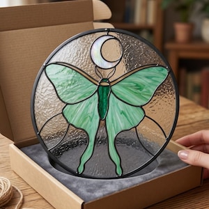Peut inclure: Un capteur de soleil en vitrail circulaire représentant un papillon lune vert et un croissant de lune blanc sur un fond en verre texturé transparent, encadré de métal noir et présenté dans une boîte d'expédition en carton ouverte.