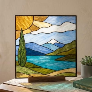 May include: A square stained glass panel depicting a landscape with a bright yellow sun, white clouds, blue mountains, a turquoise lake, and a tall green cypress tree. The panel is held upright by a rectangular wooden base stand.